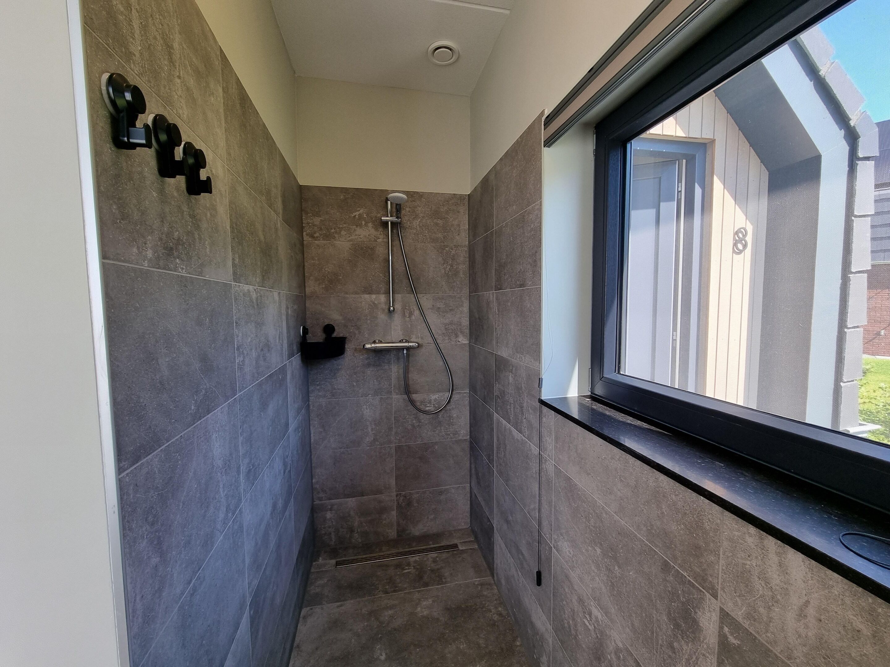 A narrow shower area with dark tiles, a handheld showerhead, and a window with a view of a building outside.