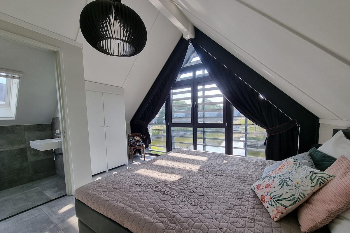 A bedroom with a large bed, a window with curtains, a hanging light fixture, and a view of the outdoors.