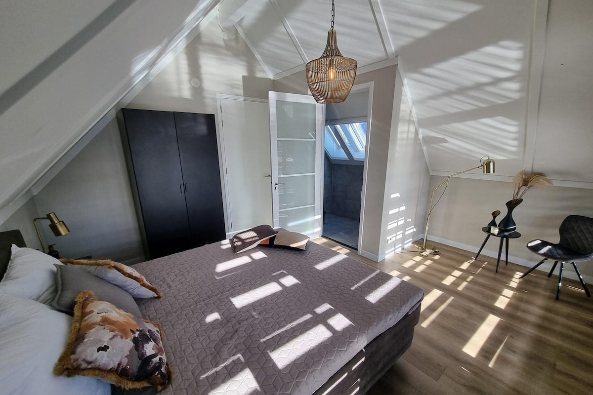 A bedroom with a sloped ceiling, a bed with a patterned bedspread, a dark wardrobe, a door leading to a balcony, and a floor lamp.