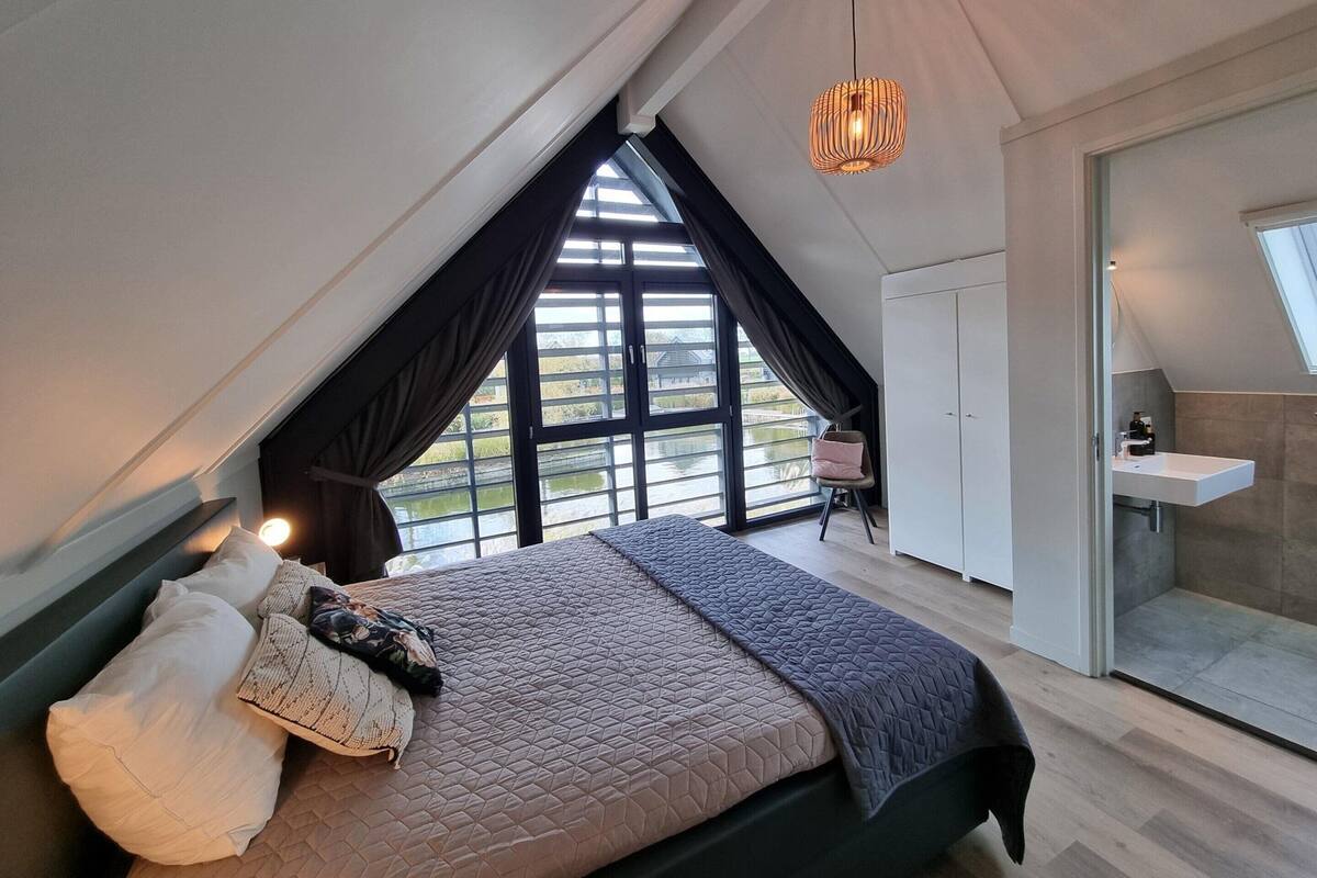A bedroom with a sloped ceiling, a bed with multiple pillows, a hanging light fixture, a chair, and a view of a body of water through large windows.
