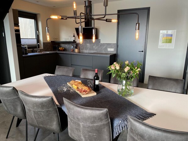 A modern dining area with a long table, grey upholstered chairs, a decorative vase with flowers, and a bottle of wine. - Luxurious Detached Water Villa With Jetty (Balk)