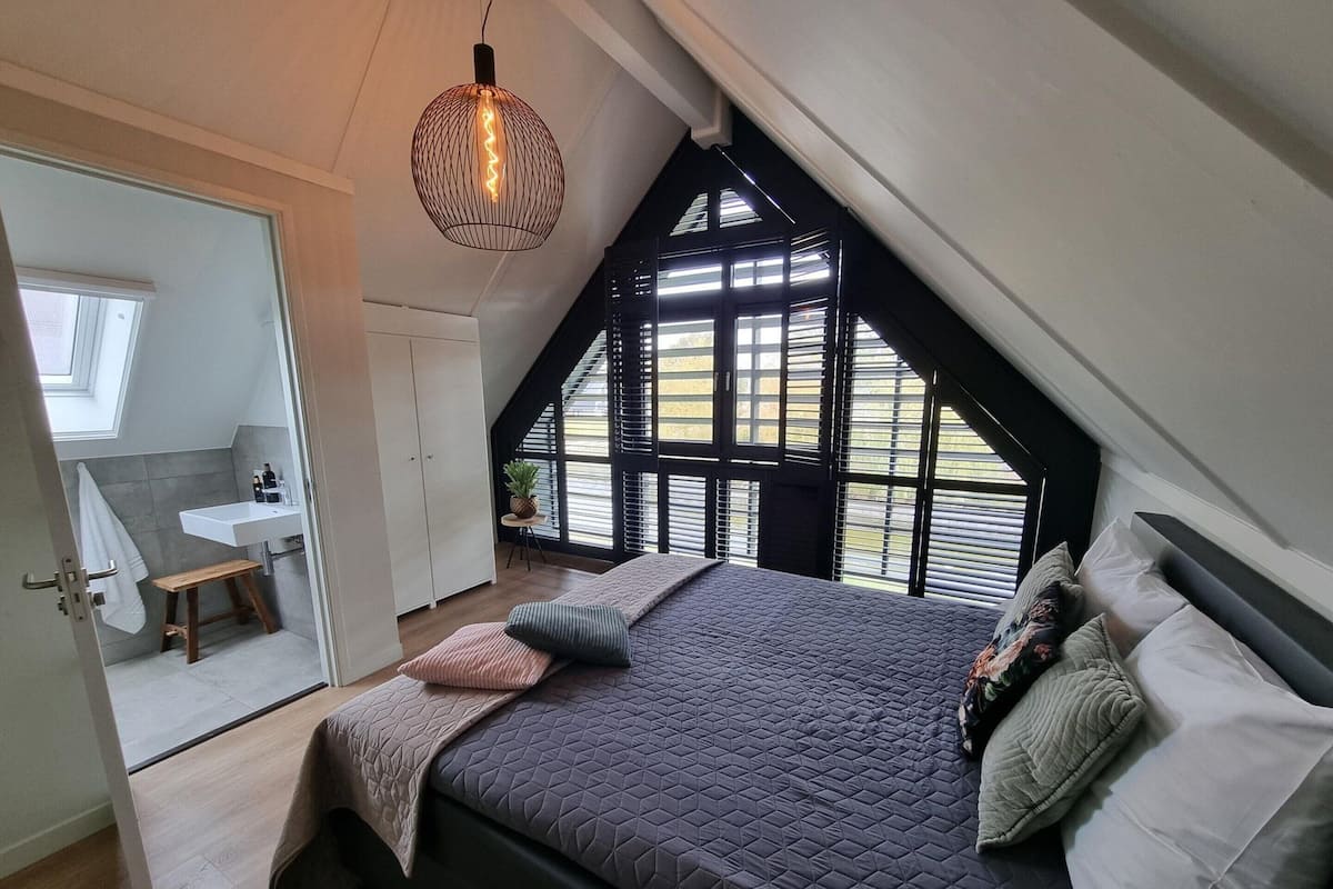 A bedroom with a sloped ceiling, a bed with multiple pillows, a bedside table, and a hanging light fixture.