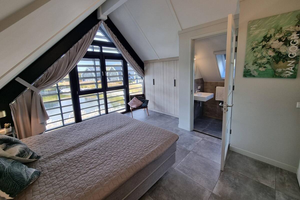 A bedroom with a large bed, a window with curtains, a chair, and a bathroom with a sink and towel rack.