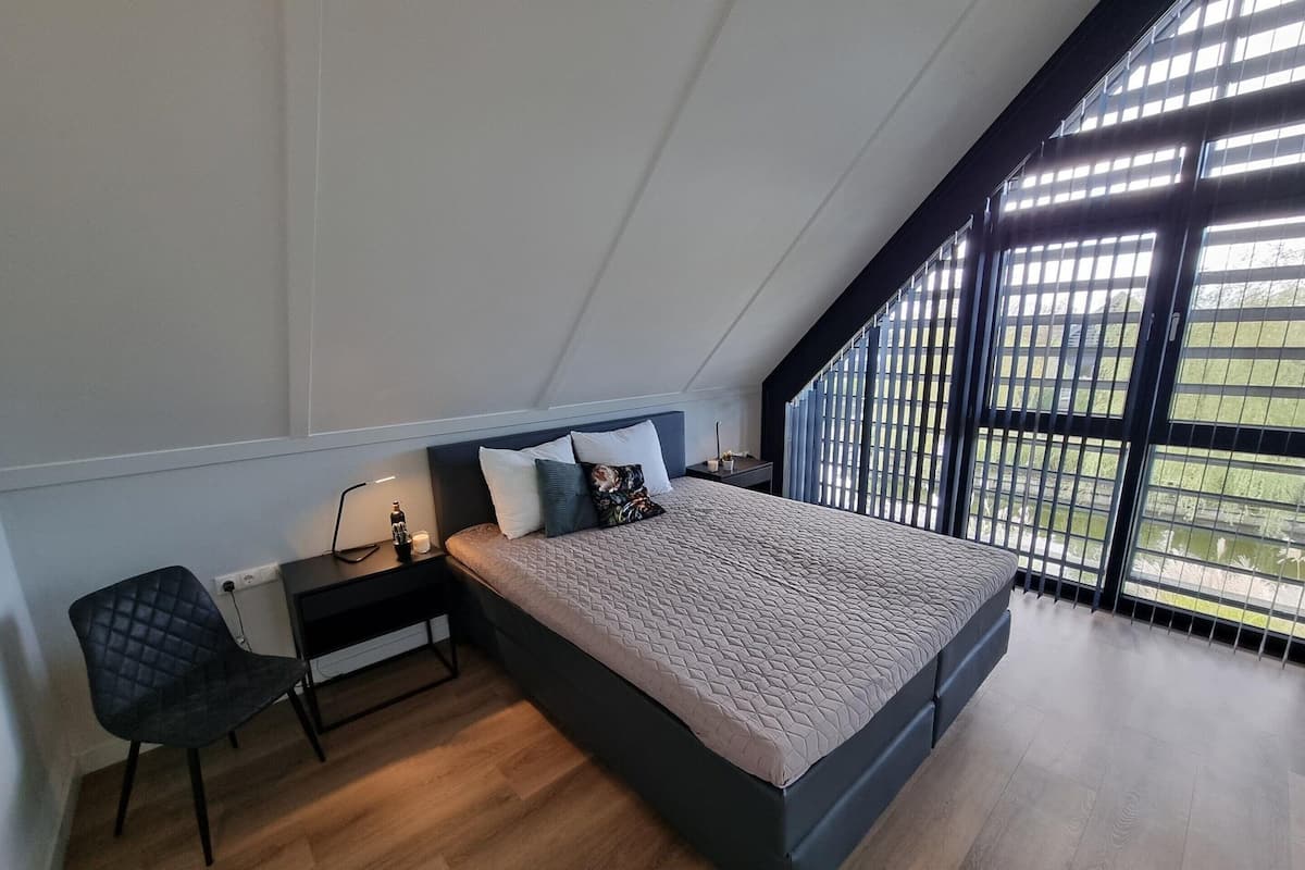 A modern bedroom with a sloped ceiling, a bed with a quilted mattress, a bedside table with a lamp, a chair, and a window with vertical blinds.