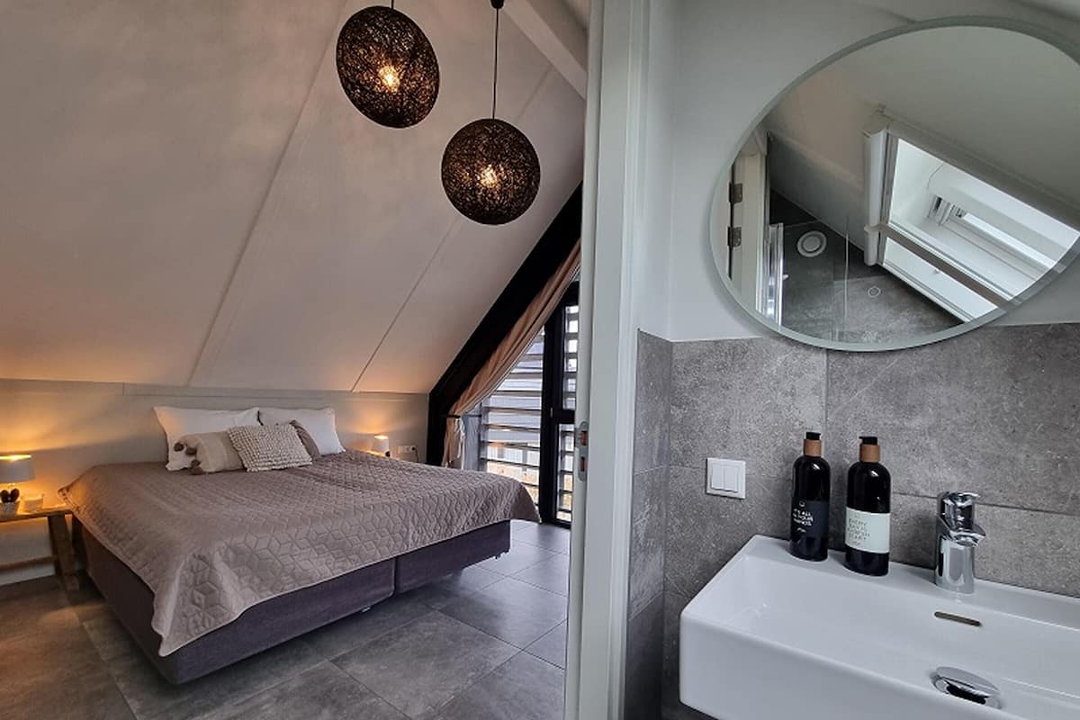 A modern bedroom with a large bed, a bedside table, a mirror, and a bathroom with a sink and two bottles.
