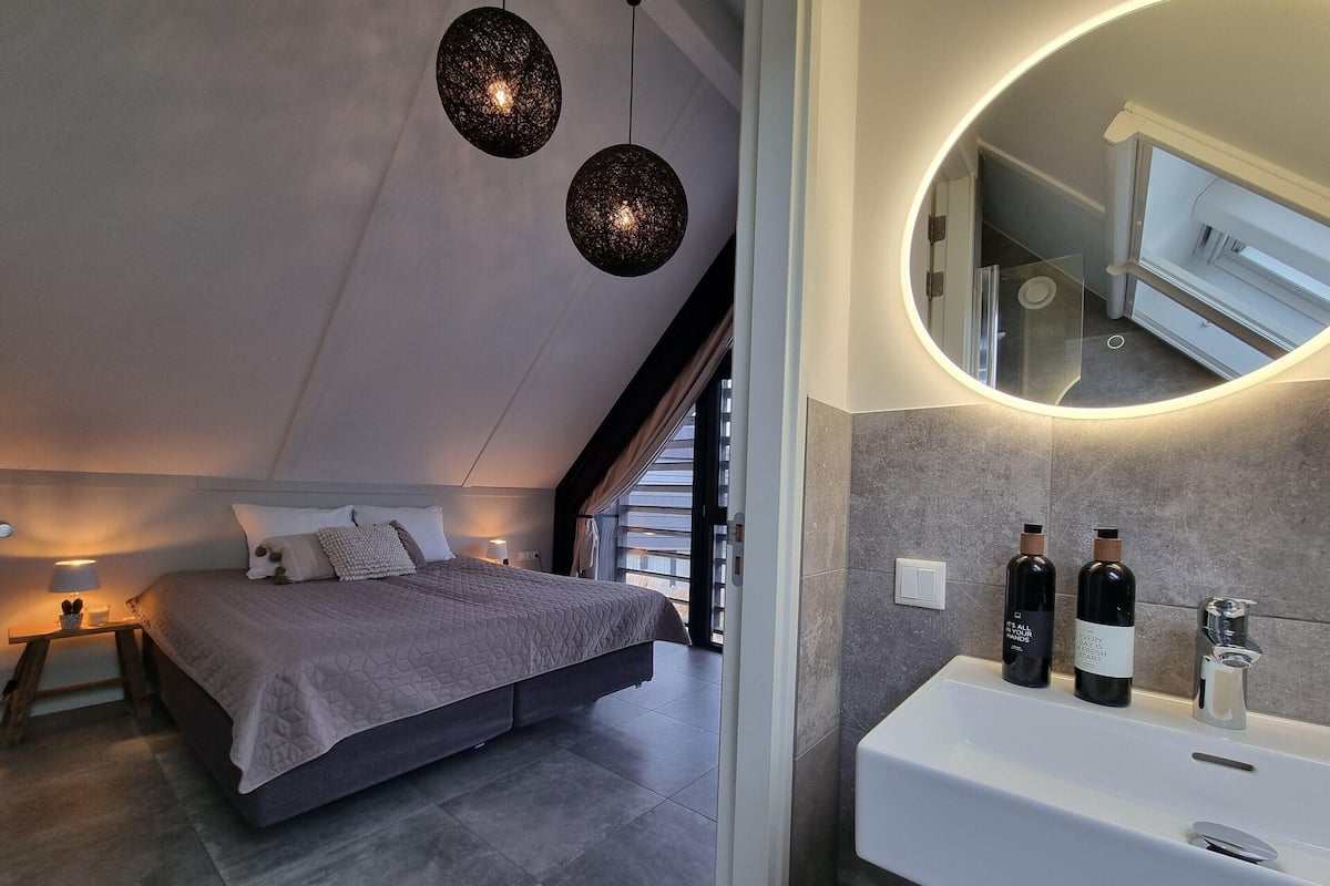 A modern bedroom with a large bed, bedside lamps, a round mirror, and a bathroom with a sink and bottles.