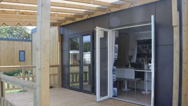 Un espace extérieur abrité doté d’une terrasse en bois et d’une porte-fenêtre coulissante donnant sur un coin cuisine avec une table et des chaises.