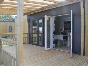 Un'area esterna coperta con un pavimento in legno, una porta scorrevole in vetro che dà su una zona cucina con un tavolo e sedie.