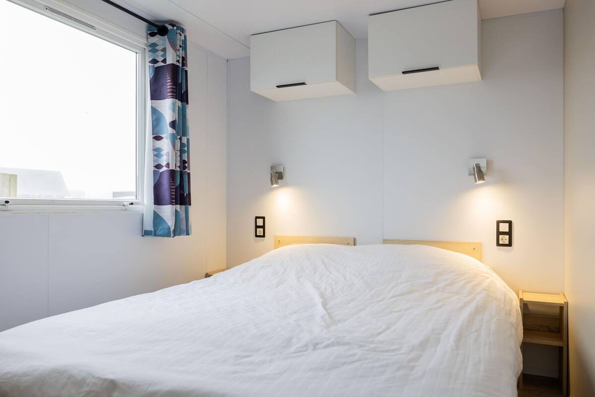 A modern bedroom with a large bed, two wall-mounted air conditioning units, bedside lamps, and a window with patterned curtains.