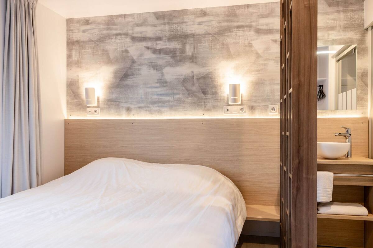 A hotel room with a large bed, a wooden headboard, a wall-mounted light fixture, a bathroom with a sink and mirror, and a wooden door.