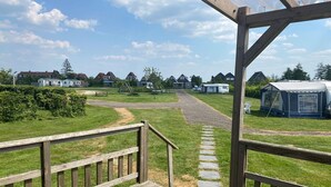 Une terrasse en bois avec un chemin de pierre menant à une zone herbeuse, où l’on aperçoit des maisons et une caravane au loin.