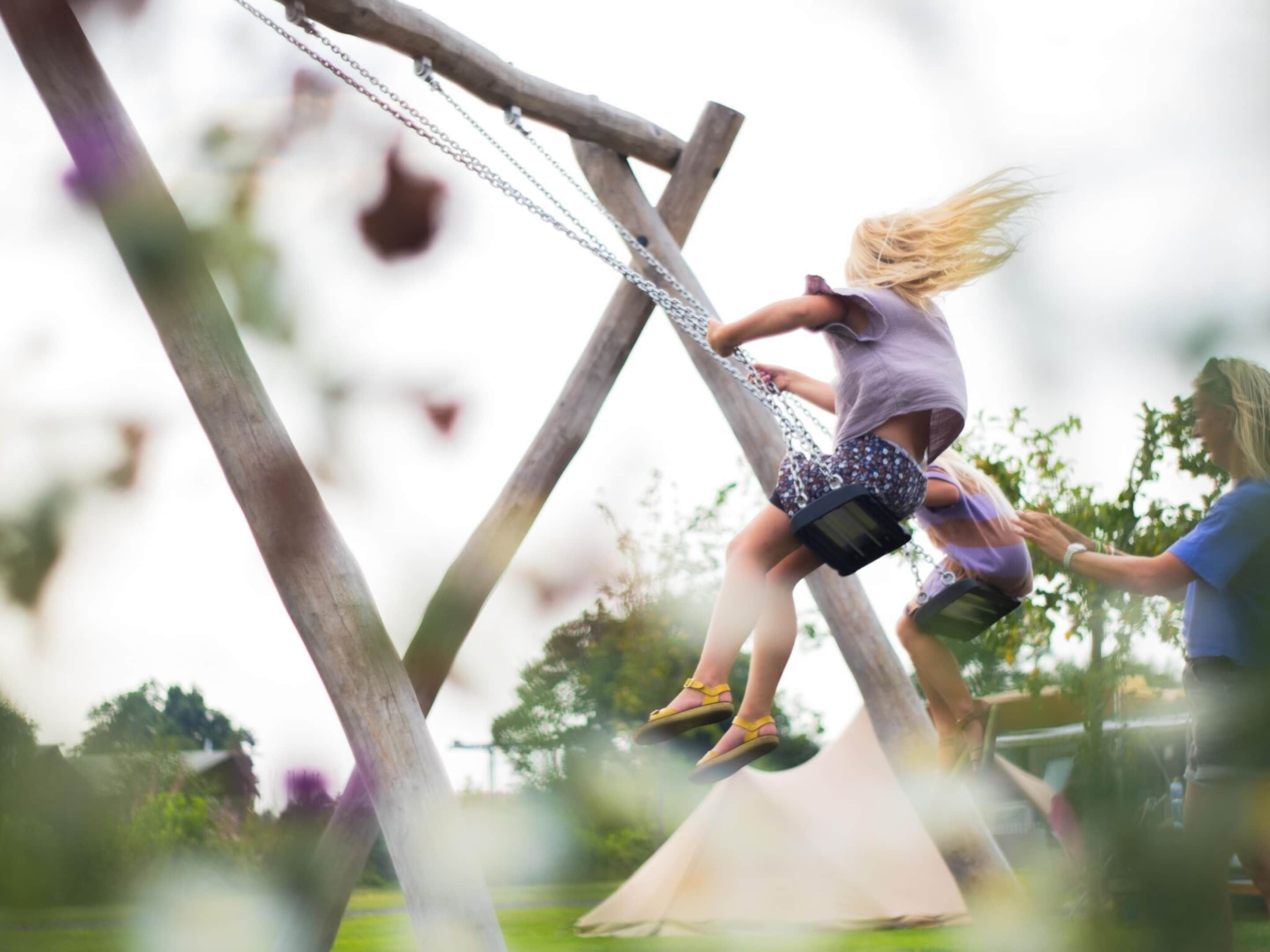 A child on a swing with a wooden frame and chains, in an outdoor setting with trees and grass.