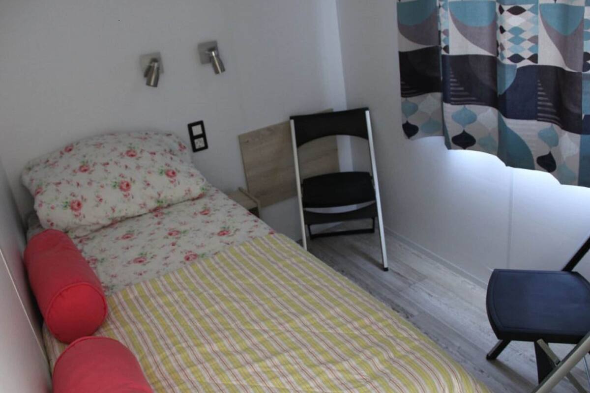 A small, neatly arranged bedroom with a single bed, a chair, and a wall-mounted shelf.