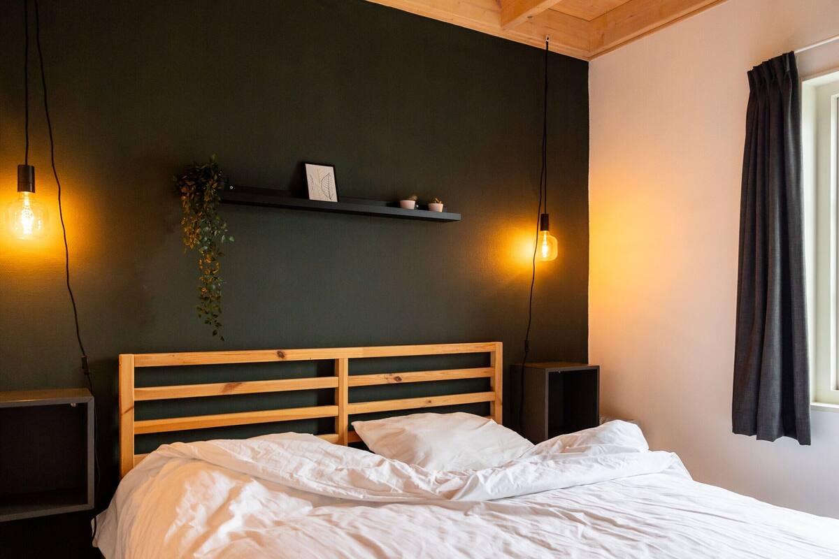 A bedroom with a wooden headboard, a bed with white bedding, two bedside tables, a wall-mounted shelf with a plant, and two hanging light bulbs.
