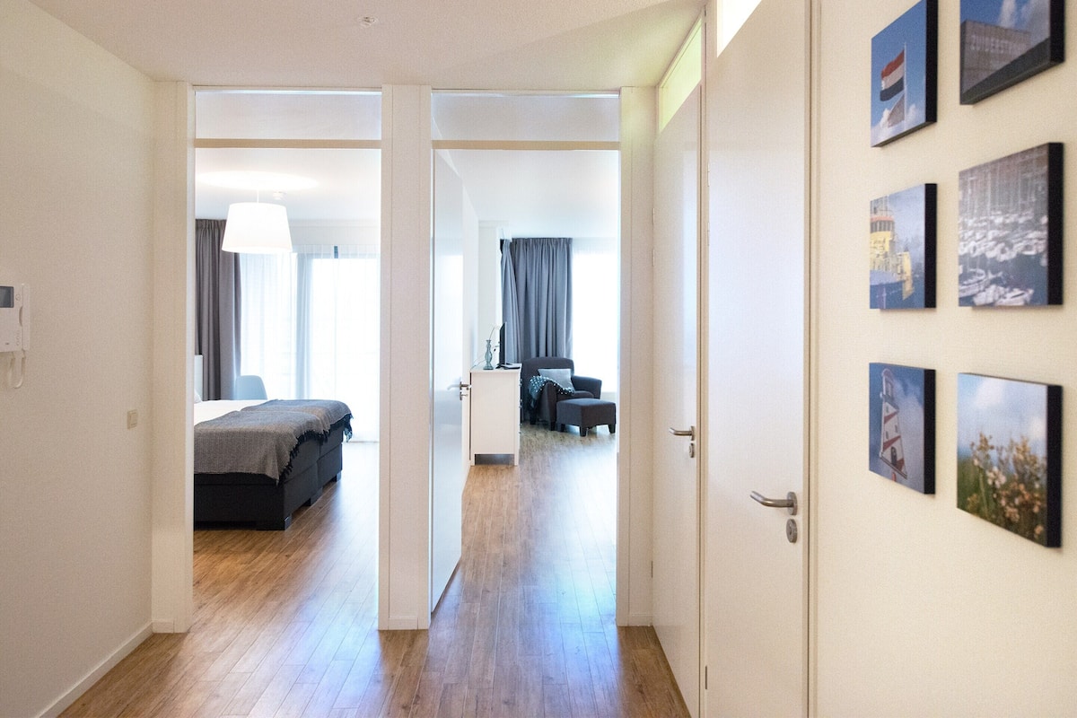 A hotel room with a bed, a desk, and a chair. There is a hallway with a door leading to another room.