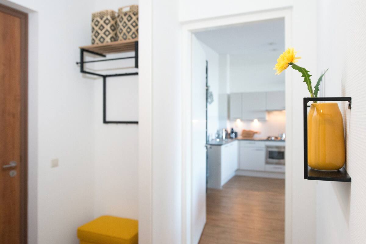 A hallway with a wooden door, a yellow ottoman, a black shelf with patterned cushions, and a yellow vase with a flower on the wall.