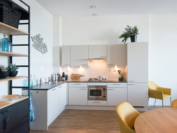 A modern kitchen with white cabinets, a microwave, and a sink. There are two yellow chairs and a wooden table. - Modern Apartment With a View of the Scheveningen Harbor (The Hague)