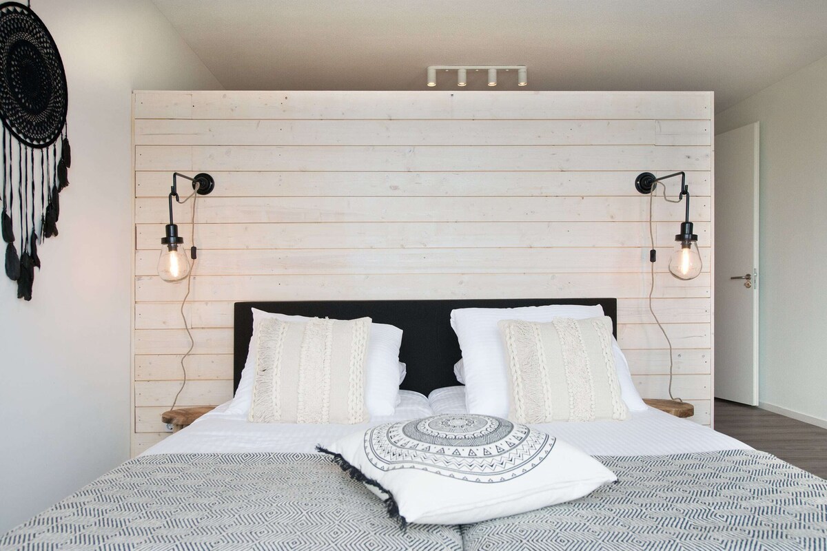 A bedroom with a wooden headboard, two bedside lamps, a patterned bedspread, and a decorative wall hanging.