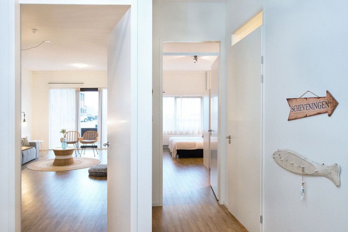 A hallway leading to a bedroom with a bed, a chair, and a small table. There is a sign on the wall that reads "Scheveningen".