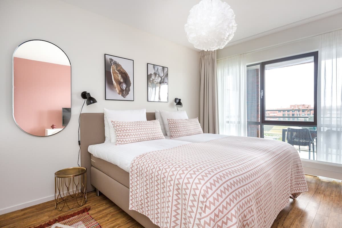 A bedroom with a bed, a round mirror, two framed pictures, a lamp, and a large window.