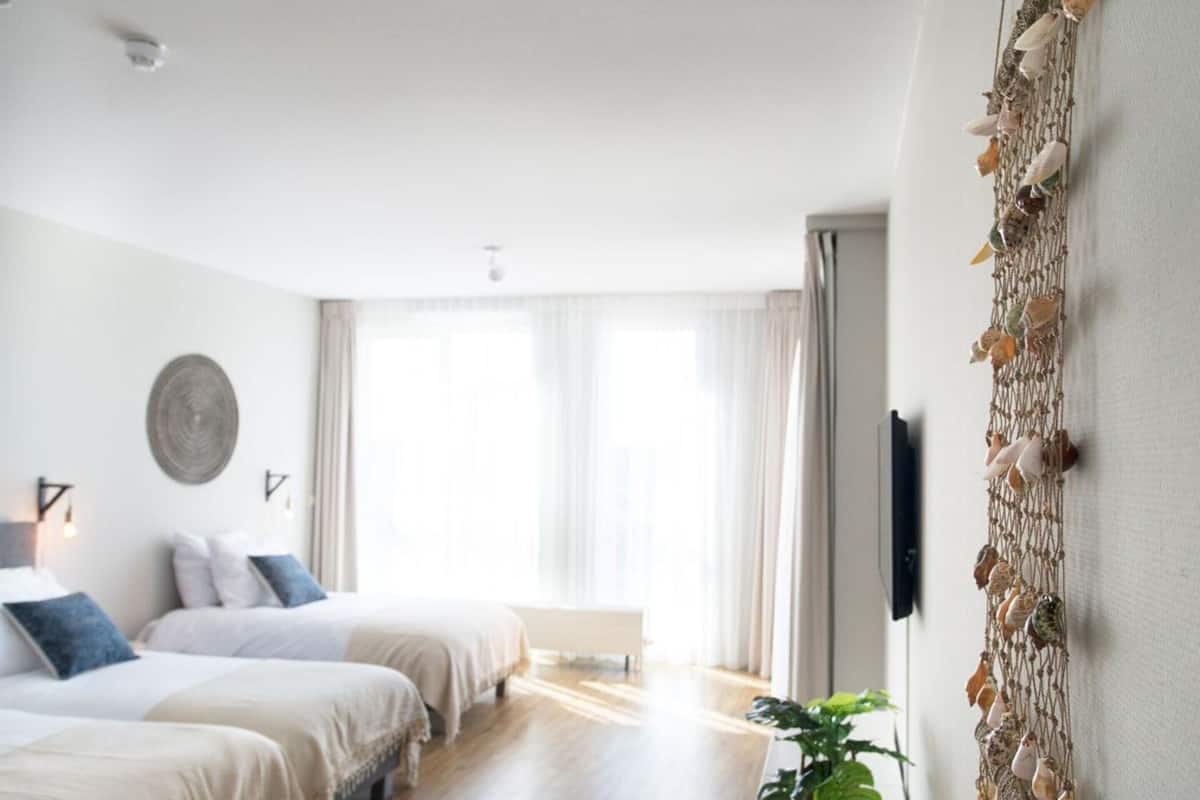A hotel room with two beds, a large window with curtains, a wall-mounted TV, and a decorative wall hanging.