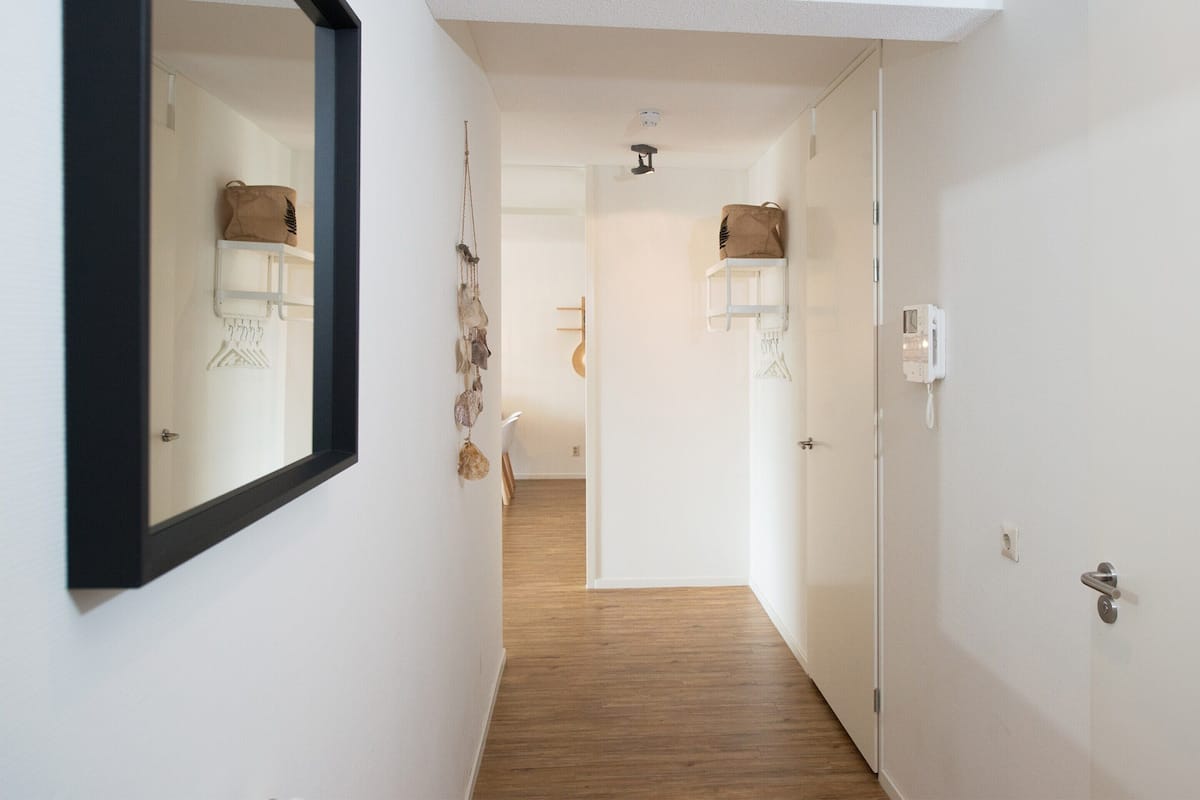 A hallway with a wooden floor, white walls, and a black-framed mirror. There are hanging items and a door with a handle.