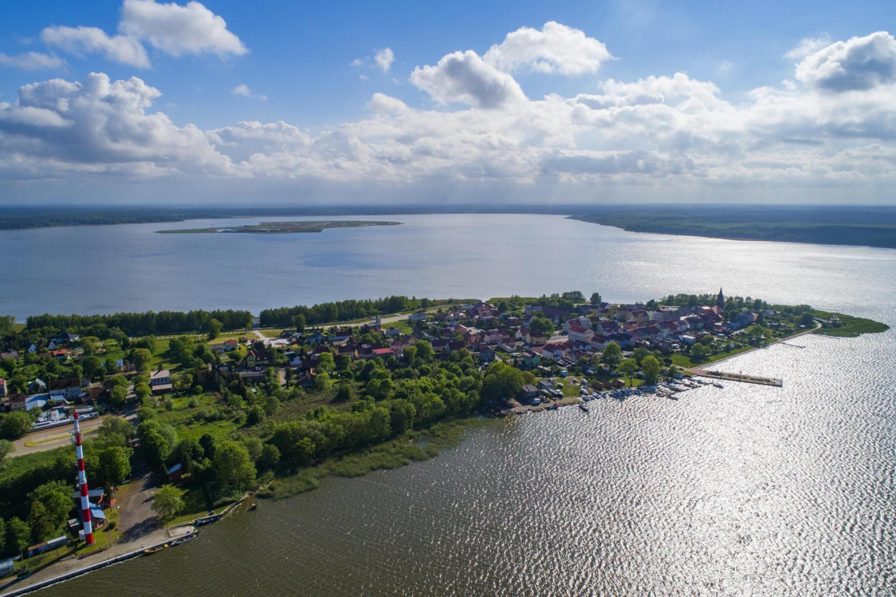 Luftaufnahme einer Stadt am Seeufer mit einer Anlegestelle, Häusern und einem Leuchtturm.