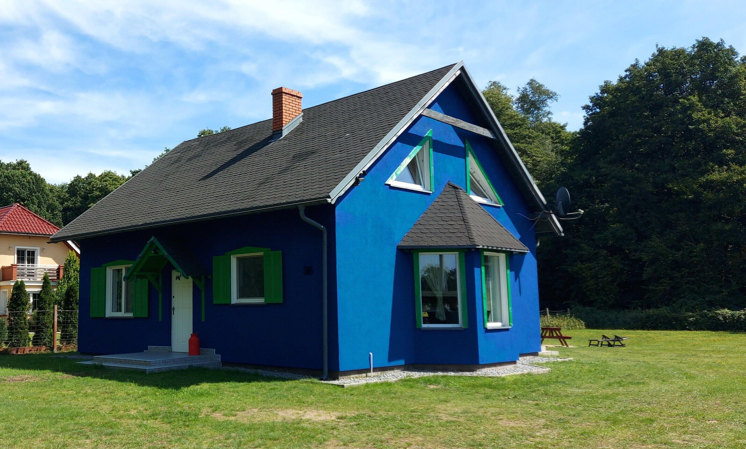A small, single-story house with a dark roof and blue exterior walls, featuring green window shutters and a chimney.