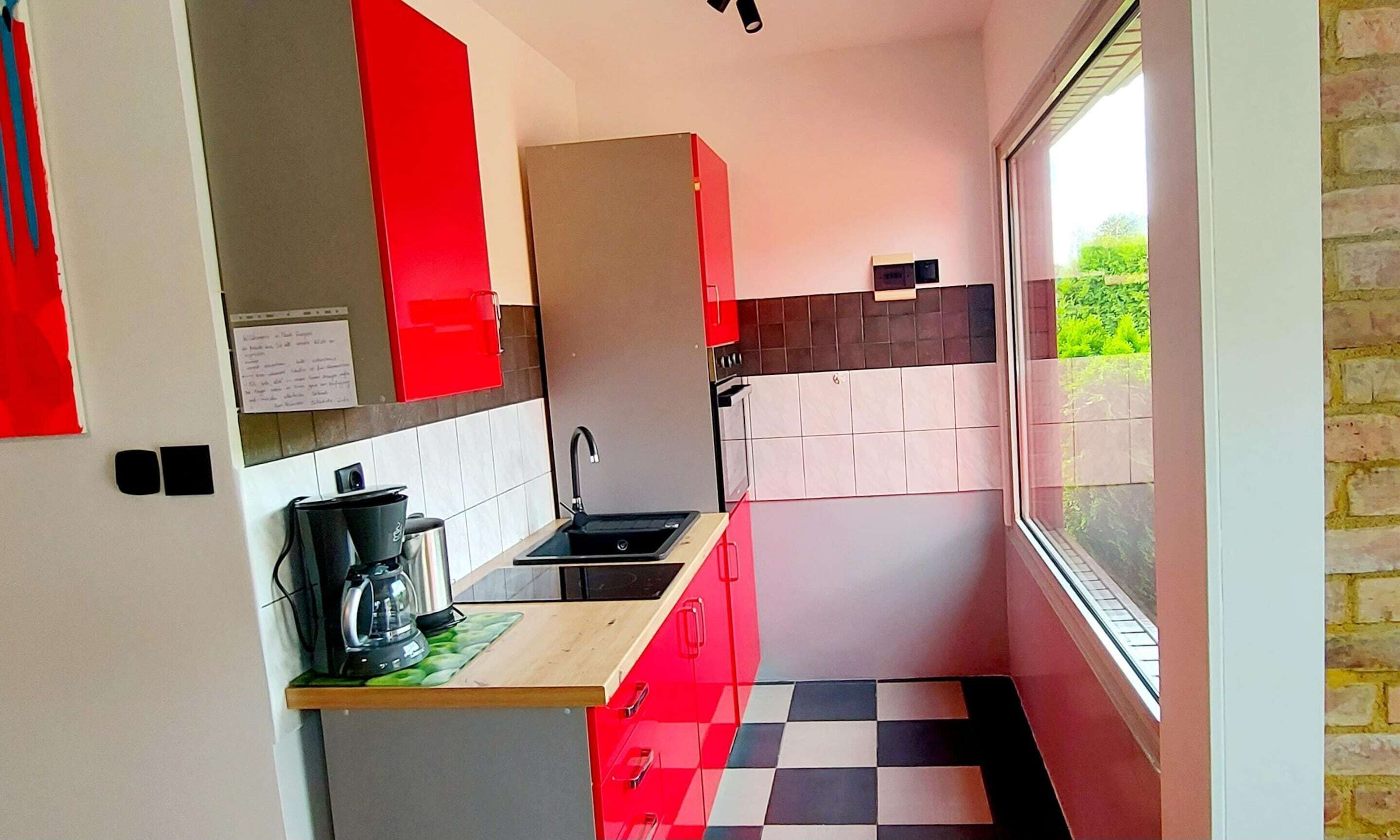 Une cuisine moderne avec des meubles rouges, un sol à carreaux noirs et blancs et une fenêtre donnant sur la verdure à l’extérieur.