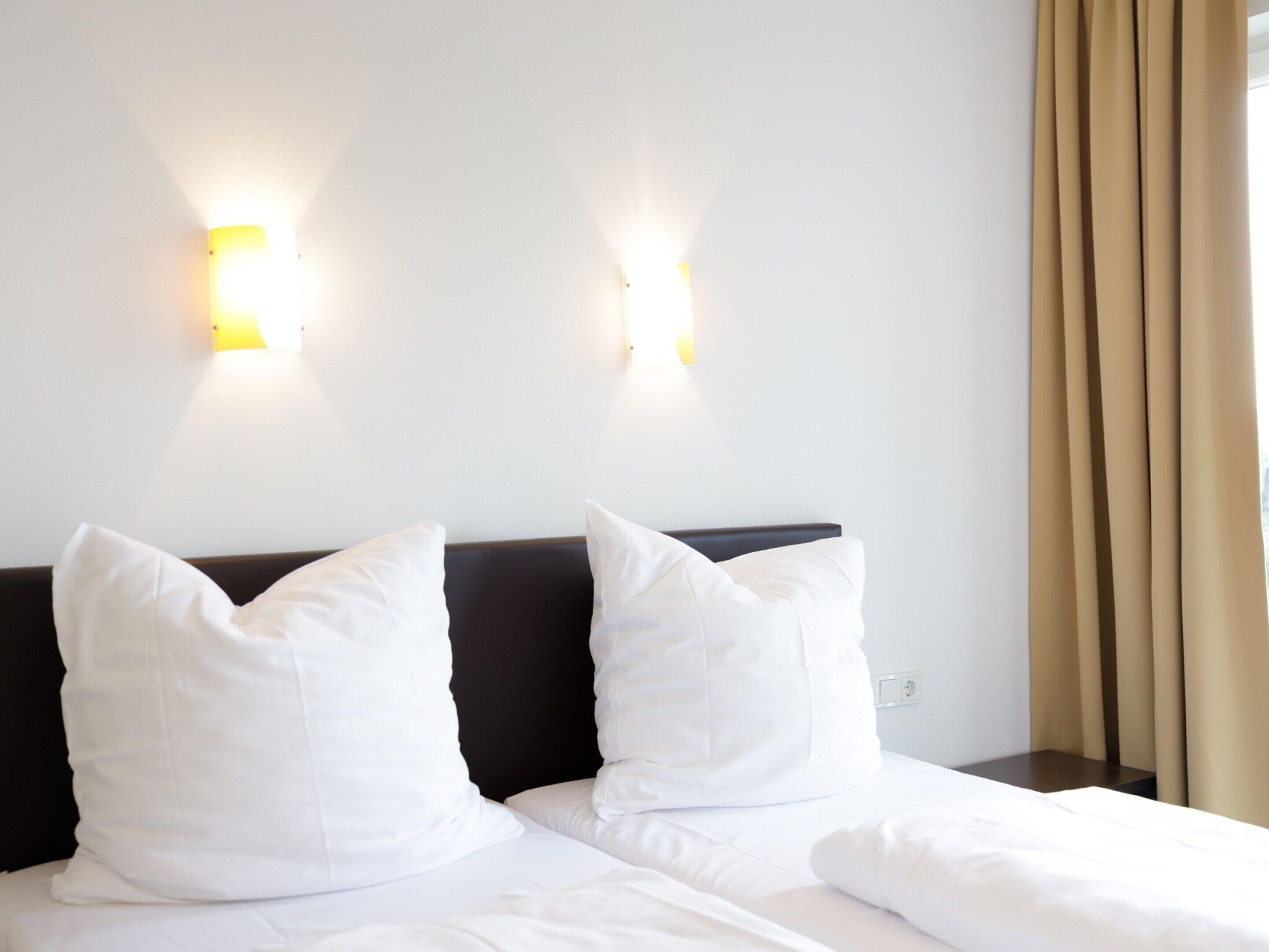 A hotel room with two beds, white bedding, a dark headboard, and wall-mounted lights.