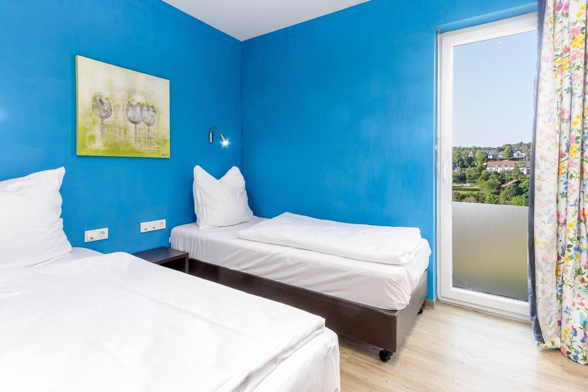 Une chambre avec deux lits, un tableau au mur, une fenêtre donnant sur un paysage et un rideau.