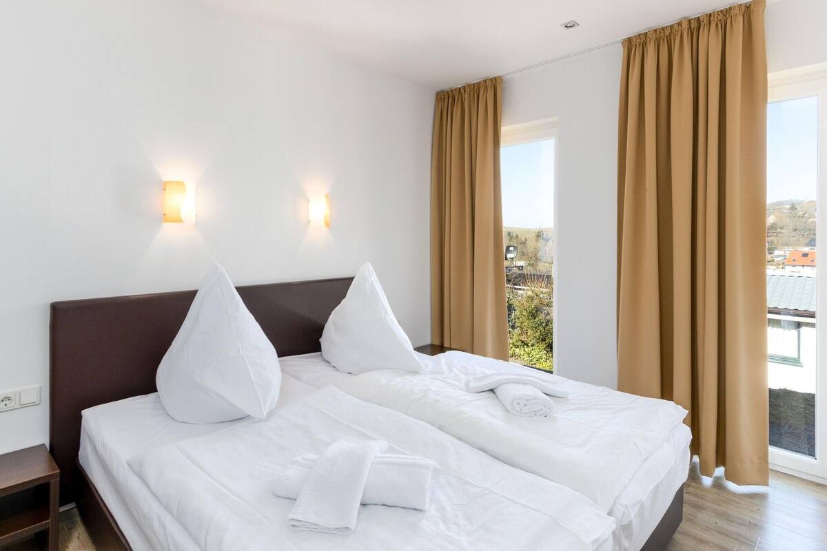 Une chambre d’hôtel avec un grand lit, deux lampes fixées au mur et une vue sur l’extérieur par une fenêtre munie de rideaux.