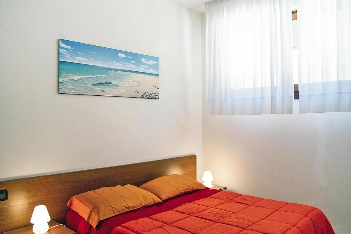 Un dormitorio con cabecera de madera, colcha roja, dos almohadas, una mesita de noche con lámpara y un cuadro de una playa.