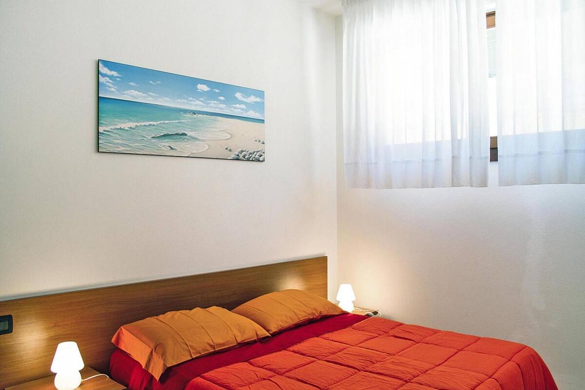 Un dormitorio con cama, mesita de noche, lámpara, un cuadro de playa y ventana con cortinas translúcidas.