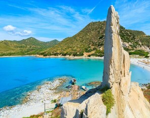 Paisagem costeira com uma formação rochosa imponente, águas azul-claras e uma área de praia com guarda-sóis.