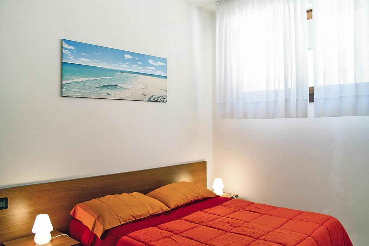 Un dormitorio con cama, mesita de noche, lámpara, un cuadro de playa y cortinas translúcidas.