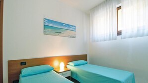 Ein Zimmer mit zwei Einzelbetten, einem Nachttisch mit Lampe, einem Bild mit Strandmotiv und einem Fenster mit durchsichtigen Vorhängen.