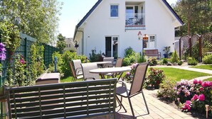 Una casa a due piani con un balcone, un patio con tavolo e sedie, e un giardino con fiori e arbusti.