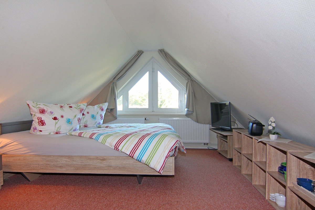 A bedroom with a bed, a TV, a window, and a shelf with dishes.