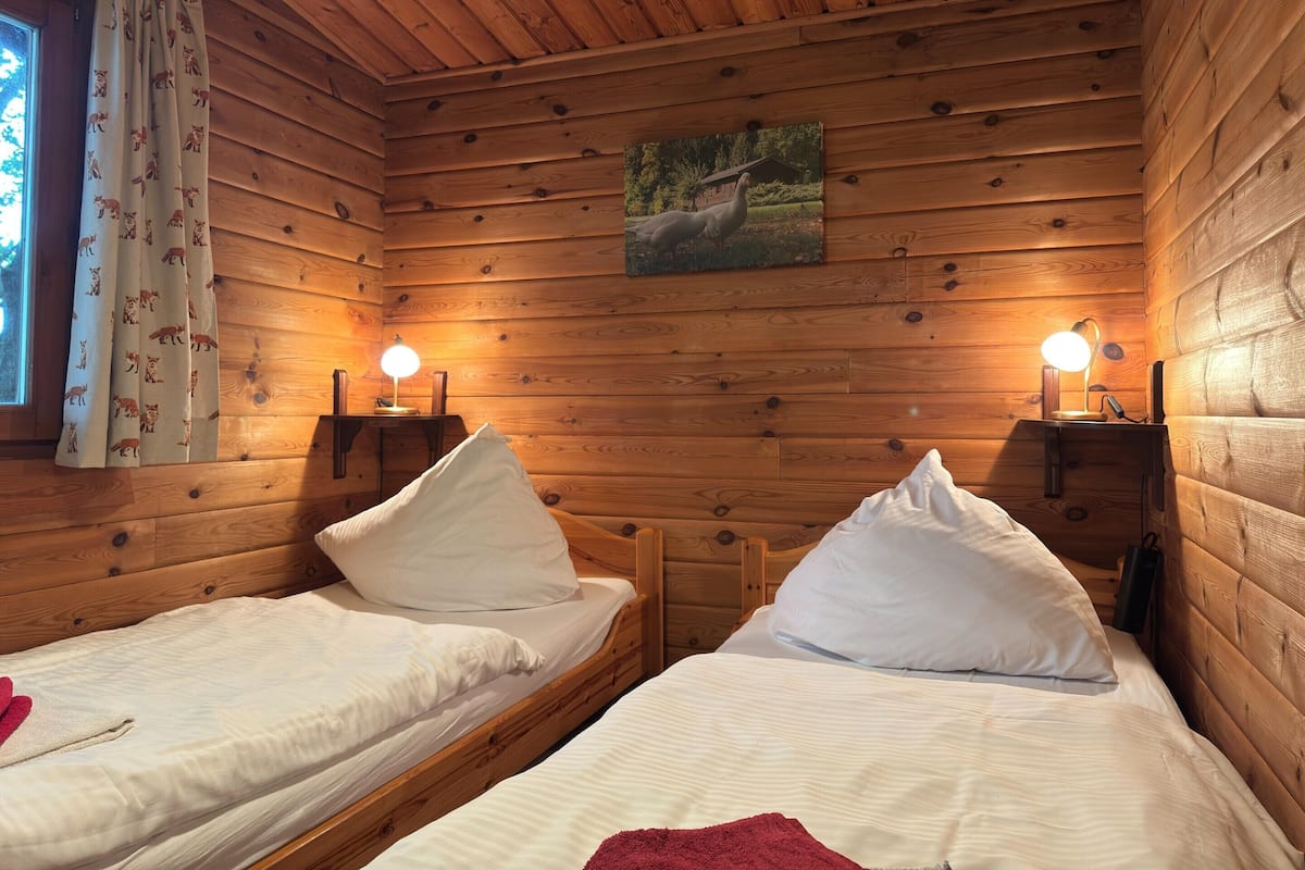 A cozy cabin room with two beds, wooden walls, a window with curtains, and a painting on the wall.