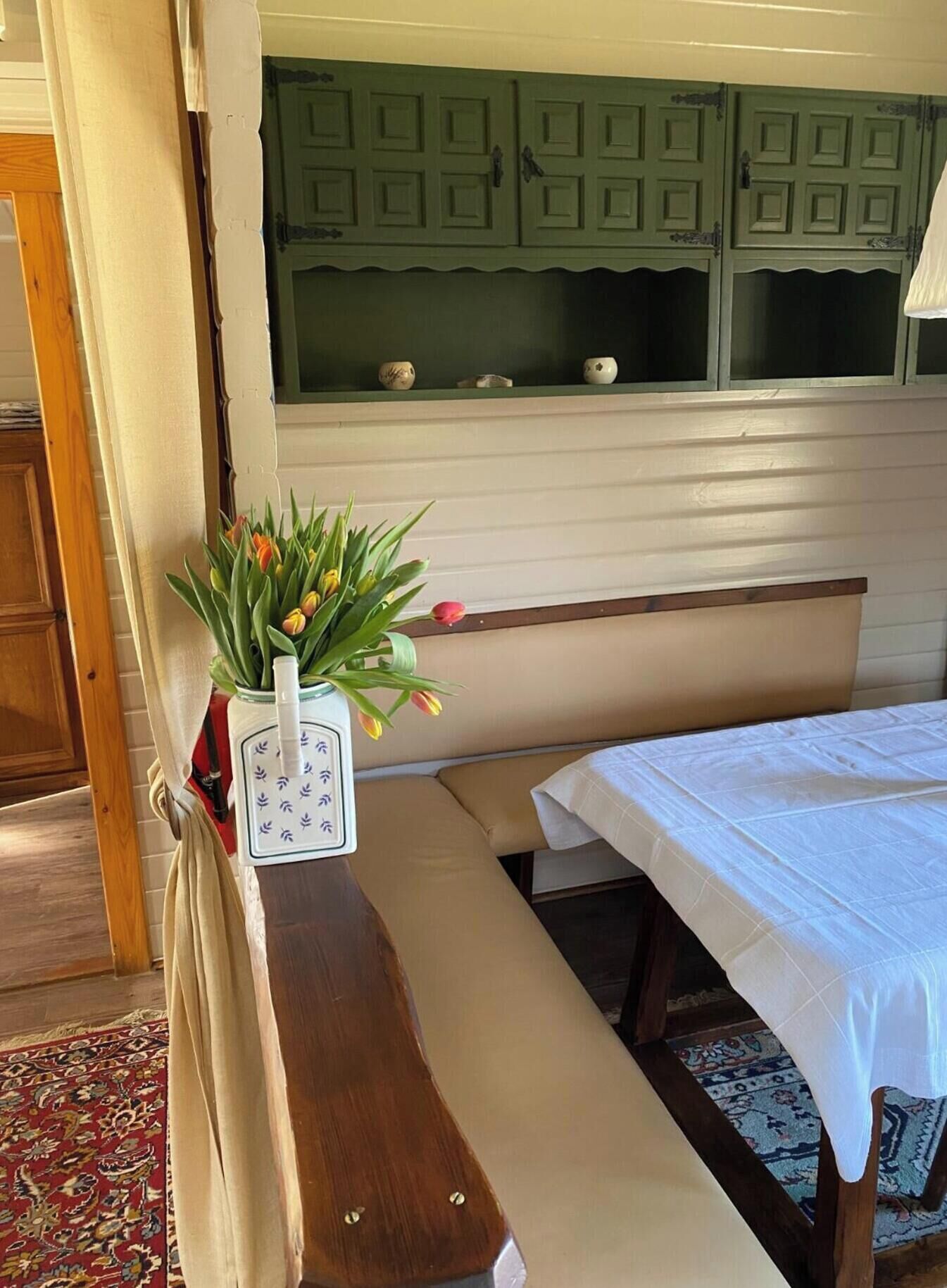 A room with a wooden bench, a small table with a white tablecloth, a potted plant with tulips, and a green cabinet with decorative items.