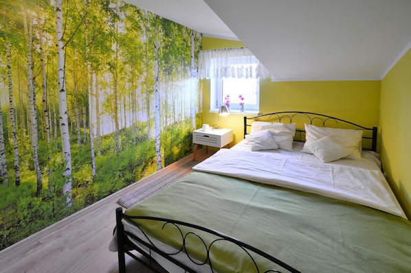 Une chambre à coucher comprenant un lit, une table de chevet, une fenêtre donnant sur des fleurs et un mur orné d’une fresque représentant une forêt.