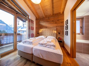 Une chambre avec un plafond en bois, un grand lit avec deux oreillers, une table de chevet et une vue sur les montagnes par la fenêtre.