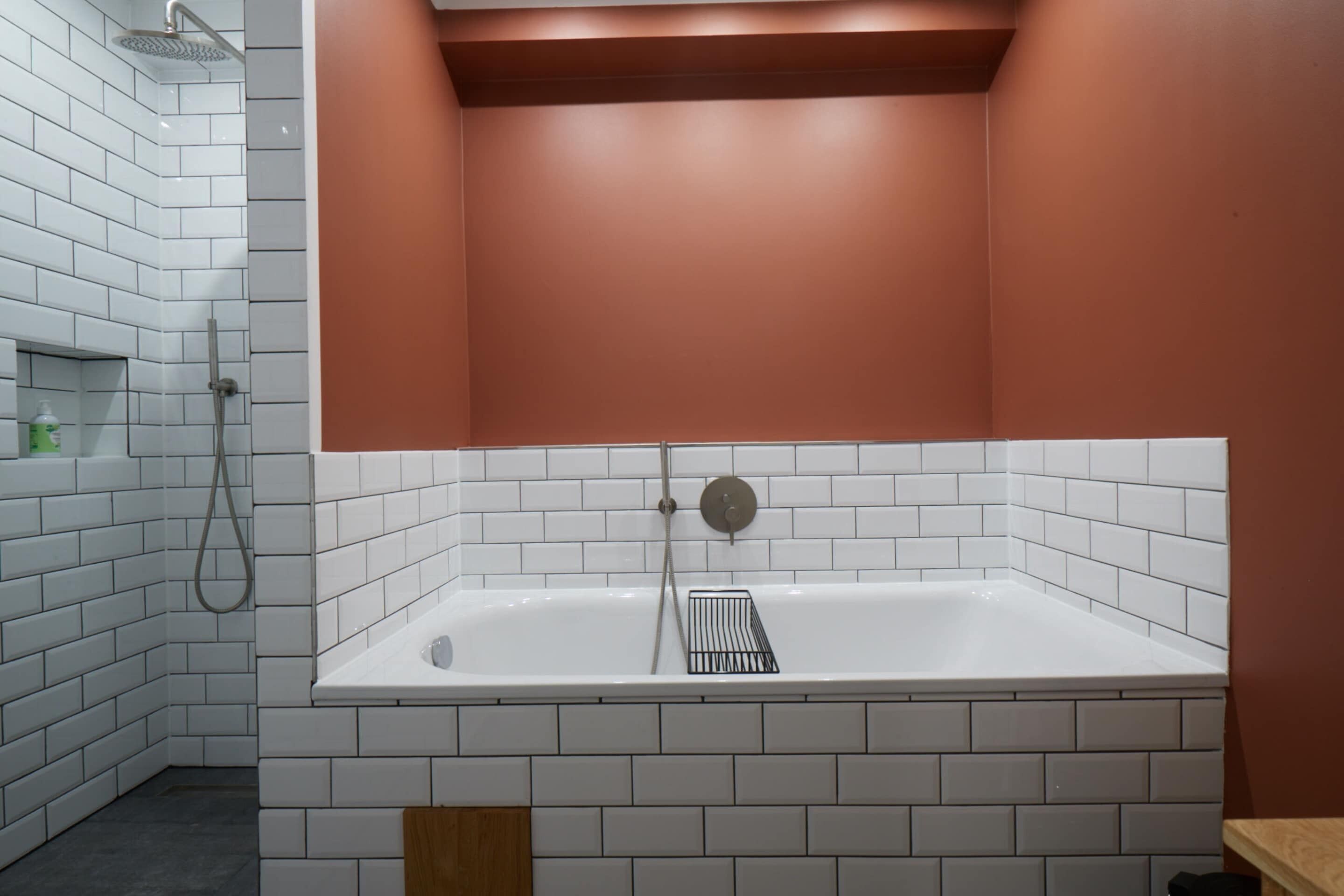 A bathroom with white tiled walls, a white bathtub, and a wooden shelf.