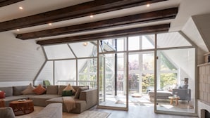 Un vaste salon doté d’une grande porte-fenêtre donnant sur un espace extérieur, de poutres en bois au plafond et d’un canapé gris agrémenté de coussins.