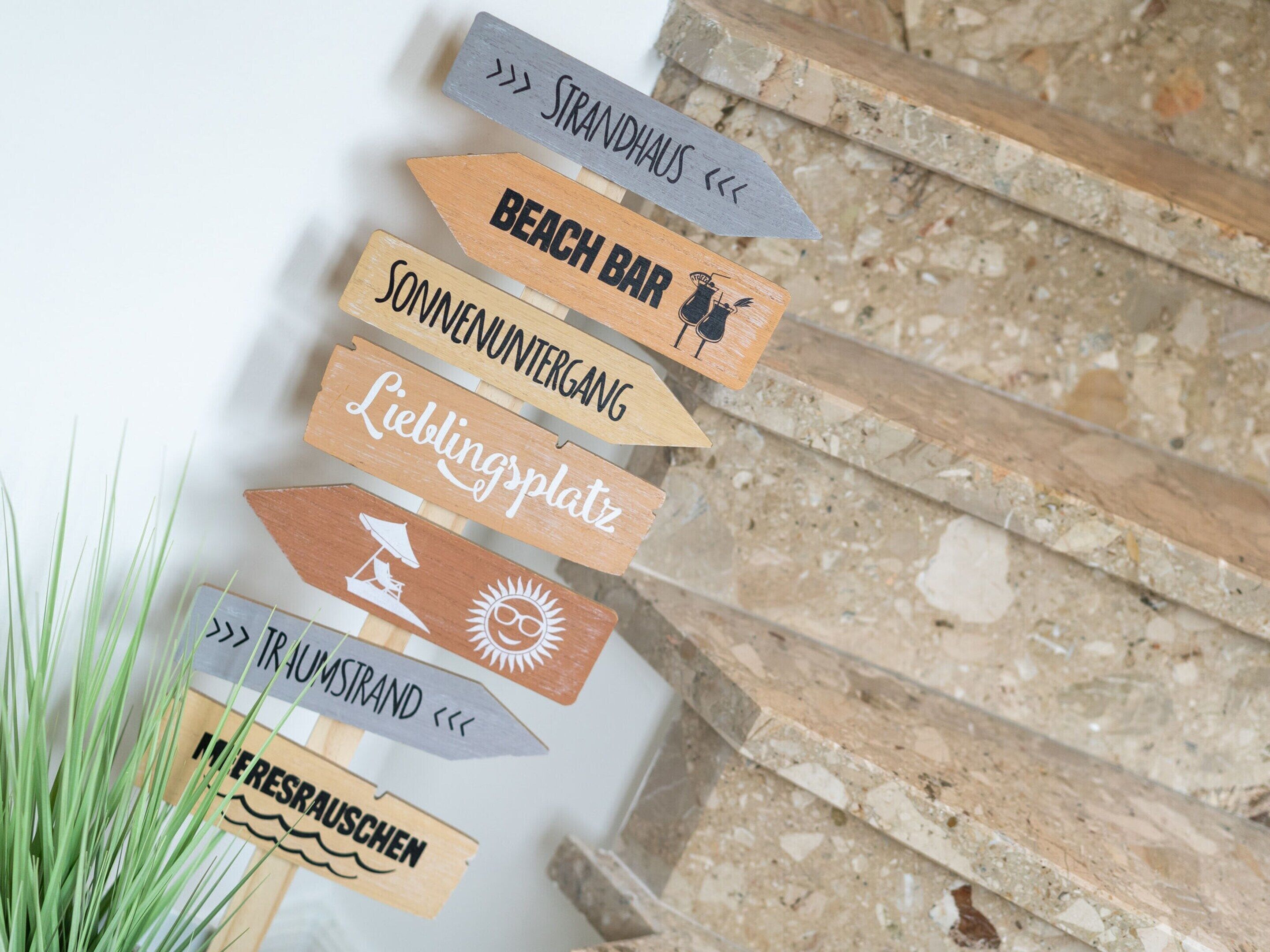 A wooden signpost with directional arrows and text indicating various locations such as 'Strandhaus', 'Beach Bar', 'Sonnenuntergang', 'Lieblingsplatz', 'Traumsstrand', and 'Frescauschen'.