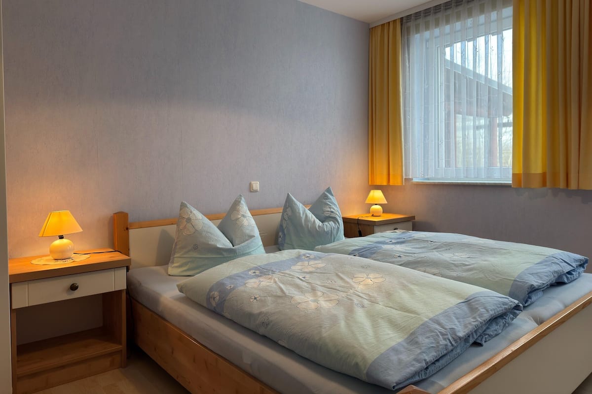 Uma cama de casal com roupa de cama florida, duas mesas de cabeceira com abajures, uma janela com cortinas amarelas e um interruptor na parede.