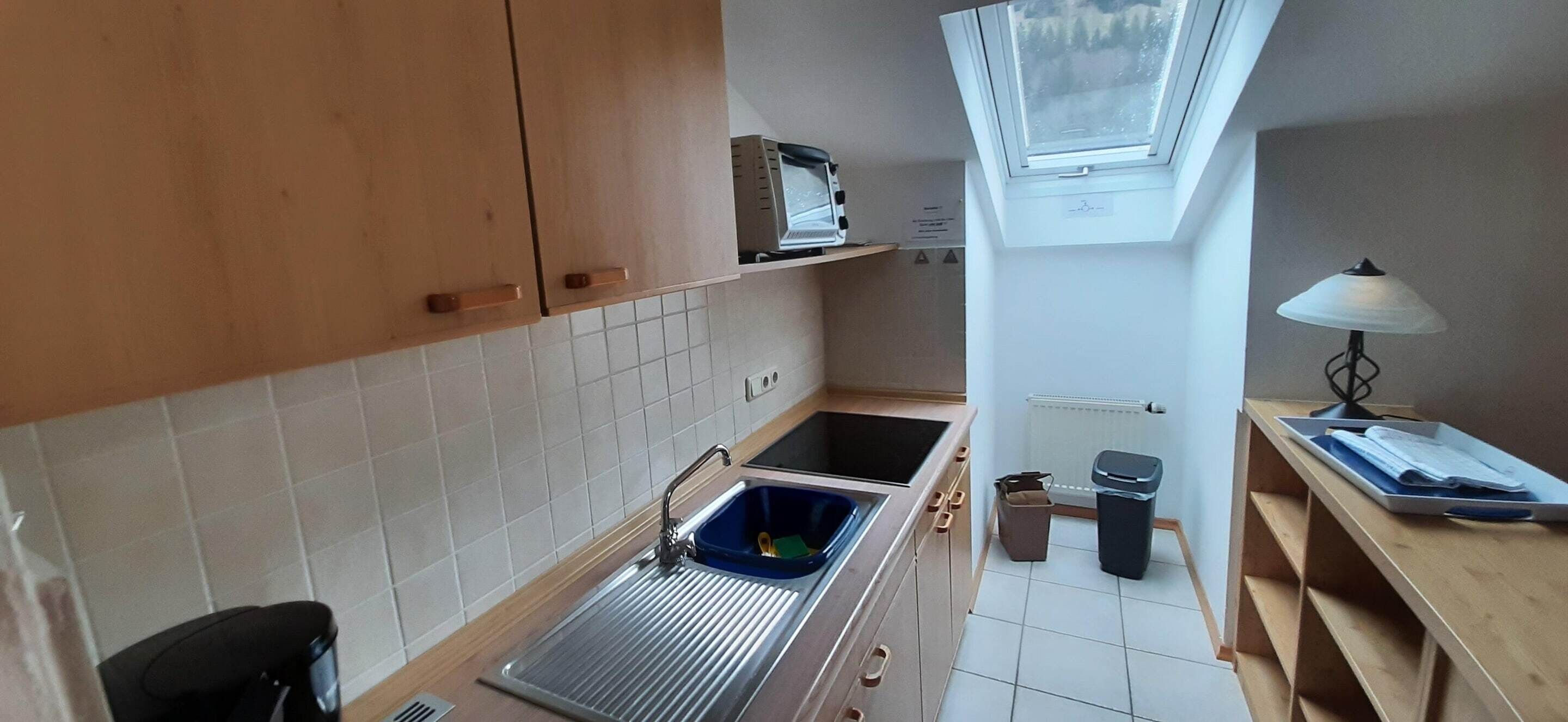 A compact kitchen with a sink, cabinets, a window, and a small table with a lamp.