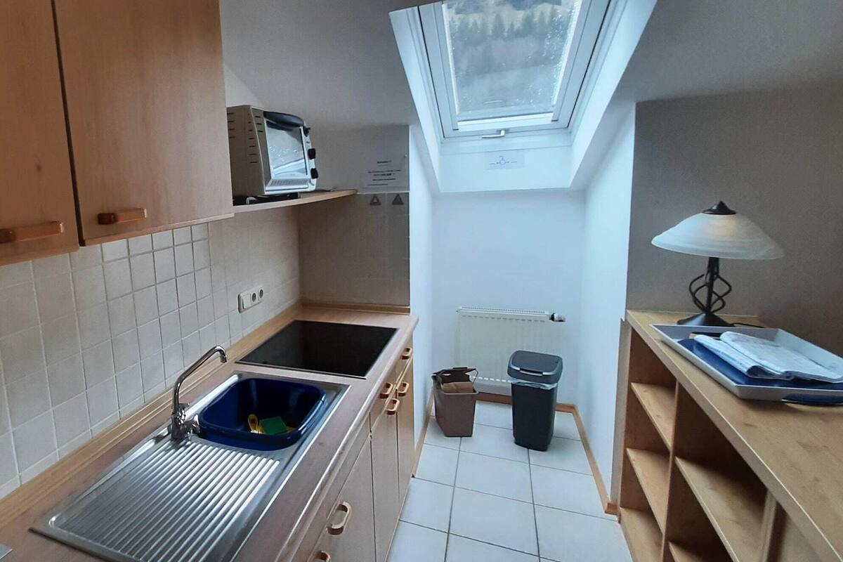 A compact kitchen with a sink, cabinets, a window, and a small table with a lamp.