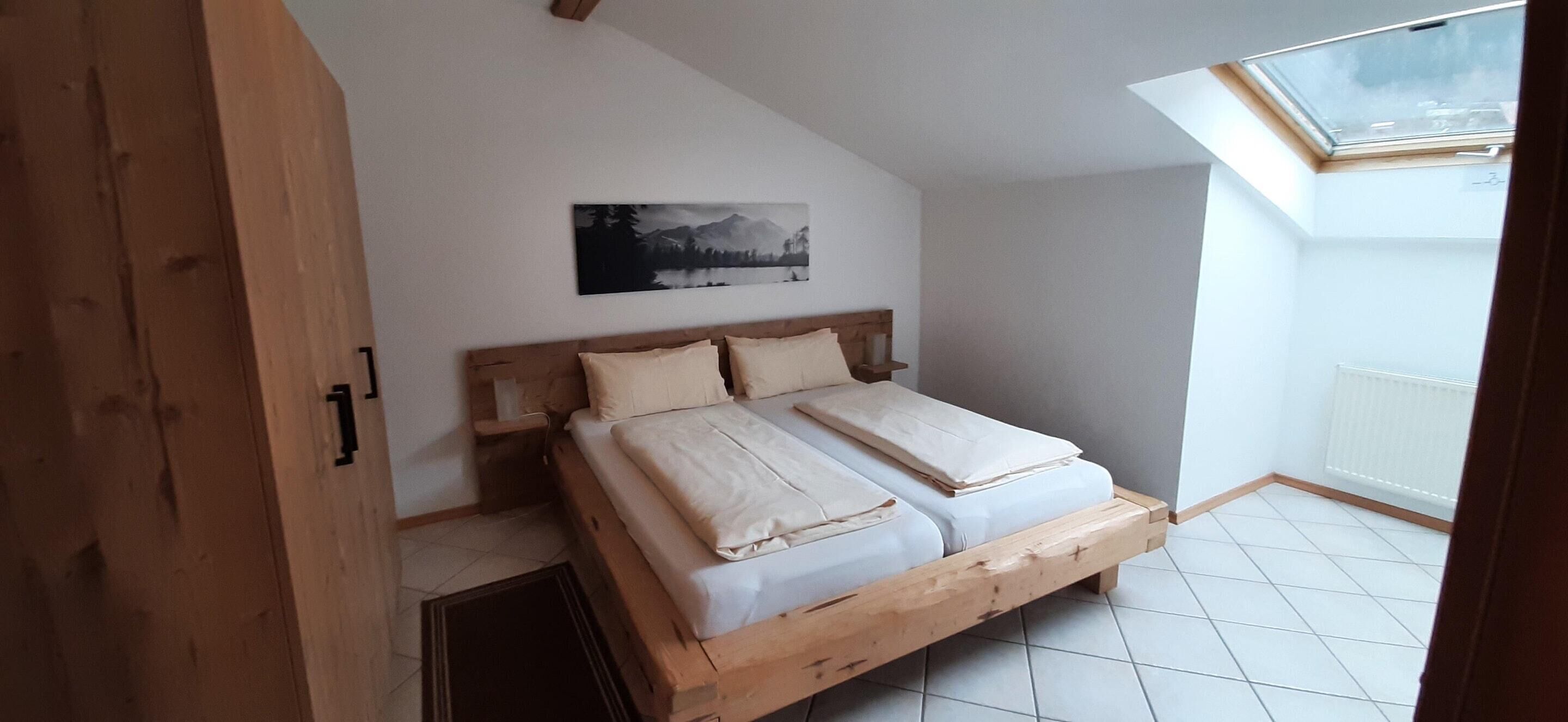 A bedroom with a wooden bed, a skylight, a white radiator, and a mountain landscape painting on the wall.